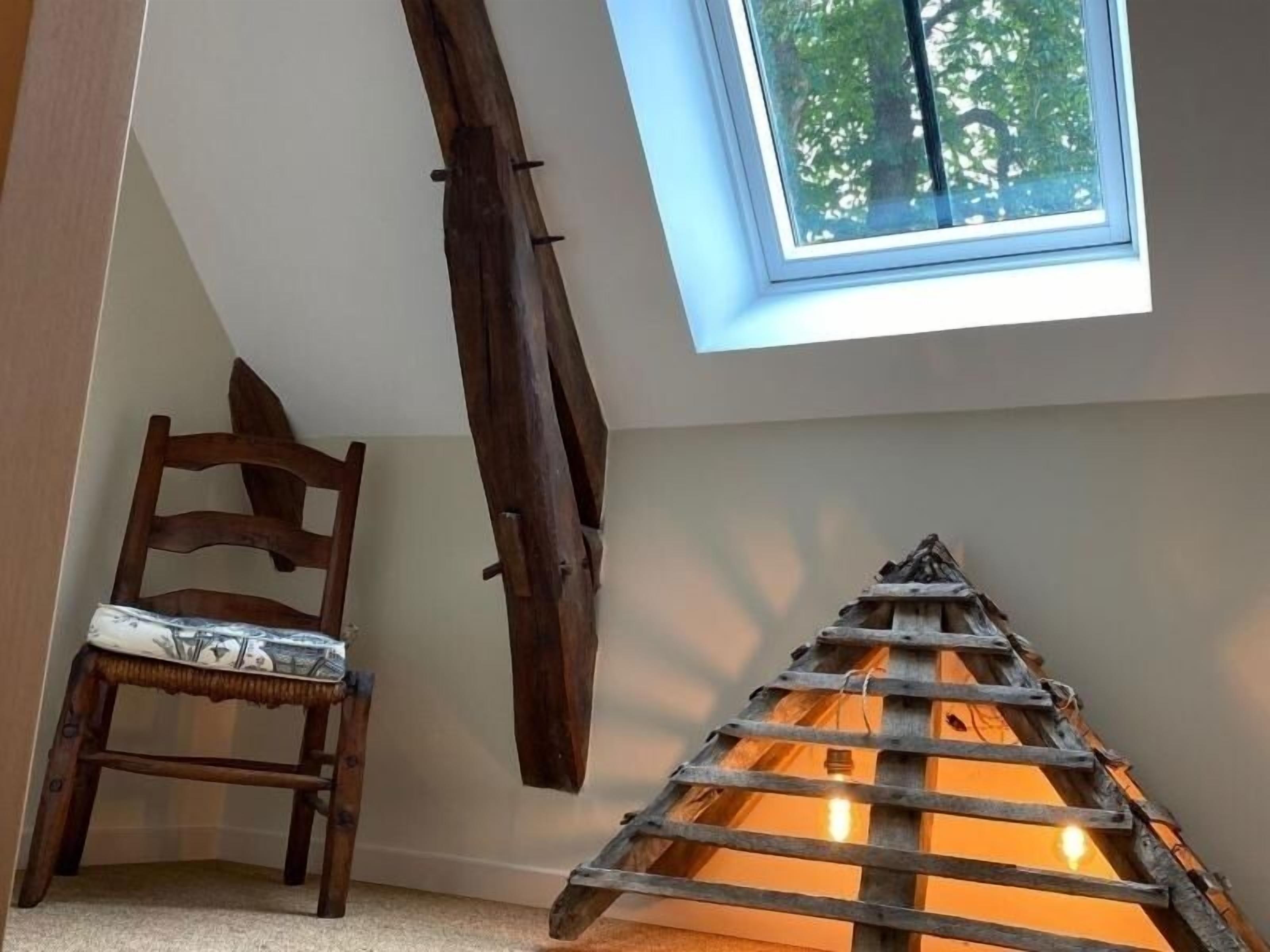 Une chambre à coucher aménagée dans un grenier, avec un plafond en pente, une lucarne, une échelle en bois et une chaise.