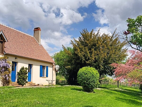 Una casa con techo de tejas rojas, contraventanas azules y un pequeño jardín con un arbusto bien cuidado y un árbol de flores rosadas.