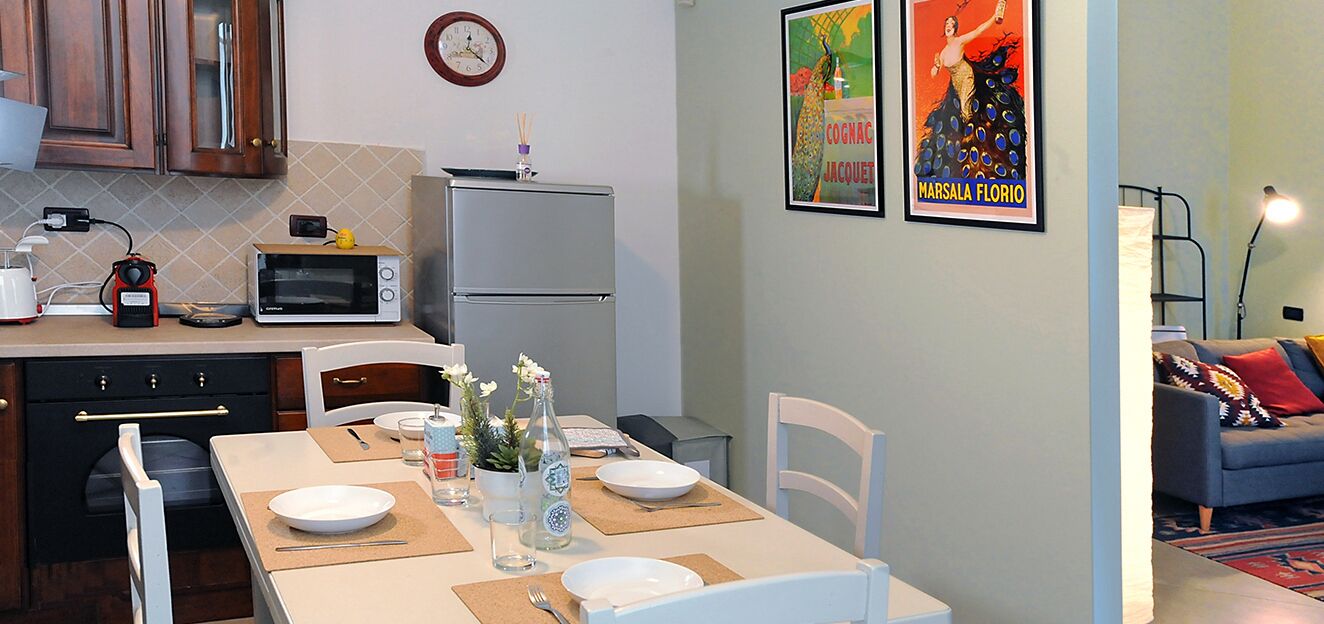 Cocina compacta con microondas, refrigerador y armarios de madera. Se observa una mesa de comedor con platos y un jarrón de flores.