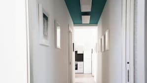 A narrow hallway with white walls, a teal ceiling, and framed pictures. It leads to a kitchen area with a refrigerator and cabinets.