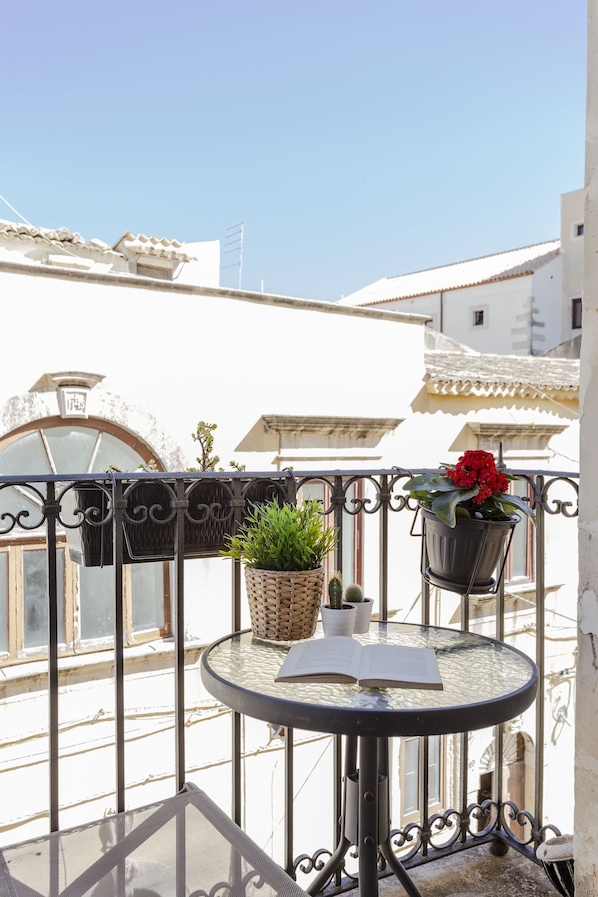 A balcony with a round table, two hanging planters with plants, and a window with a view of buildings.