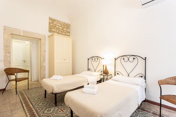Habitación de hotel con dos camas individuales, una silla, una mesita con lámpara y una pared de piedra.