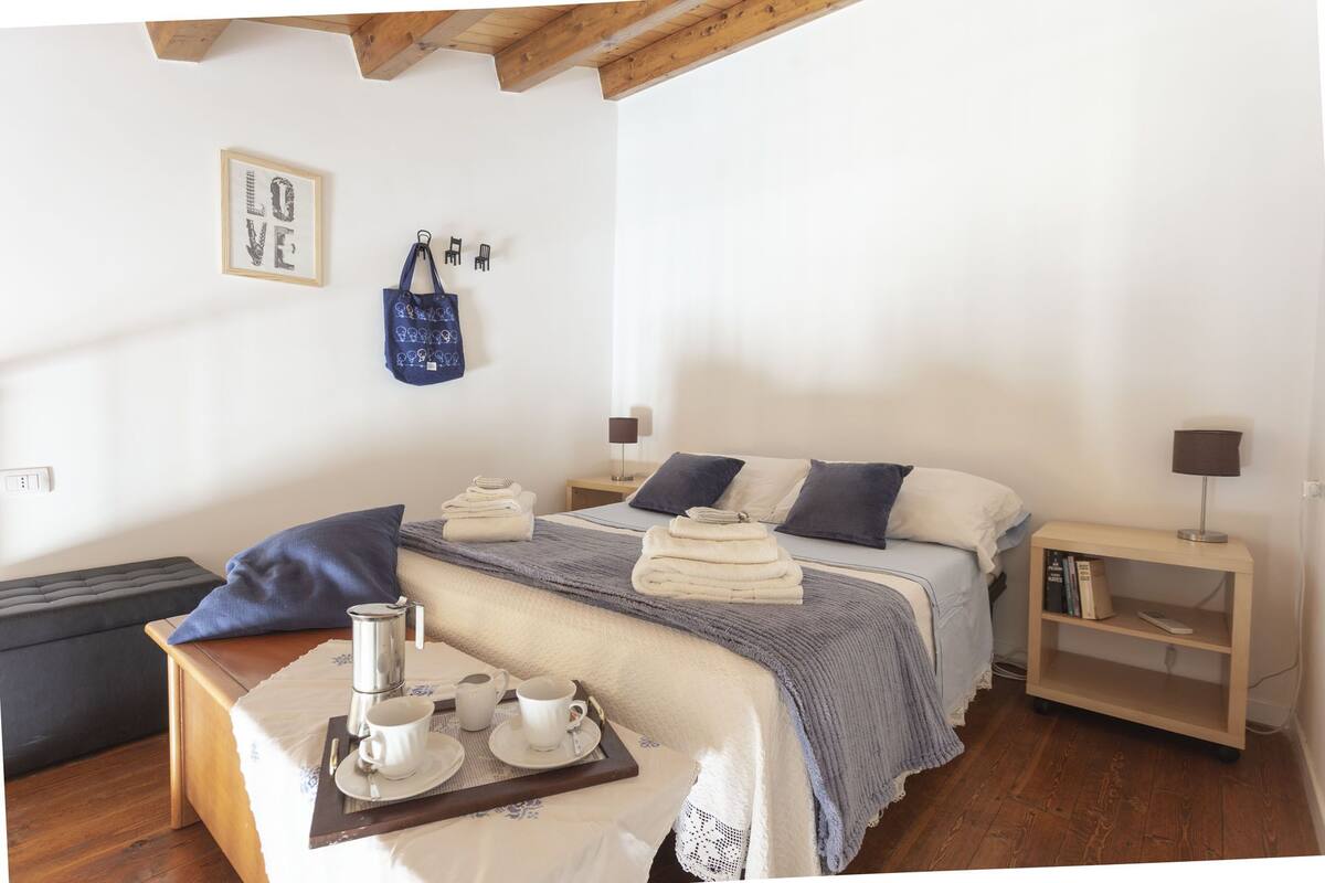 Een keurig opgemaakte slaapkamer met een bed, nachtkastjes, een zithoekje en een tas die aan de muur hangt.