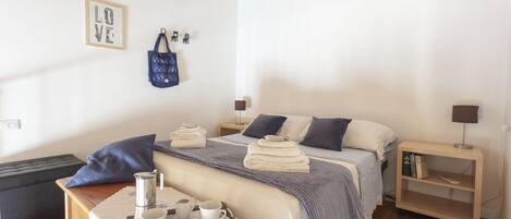 Un dormitorio ordenado con cama, mesitas de noche, un pequeño rincón de descanso y un bolso colgado en la pared.