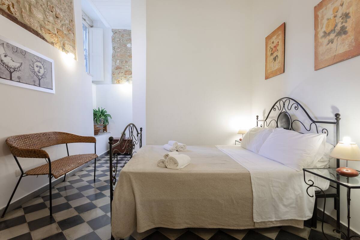 Een hotelkamer met een geruit vloerkleed, een bed met witte lakens, een hoofdeinde van metaal, een houten stoel en kunstwerken aan de muren.