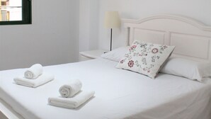 A white bedroom with a bed, a window, a wall-mounted lamp, and a ceiling light fixture.