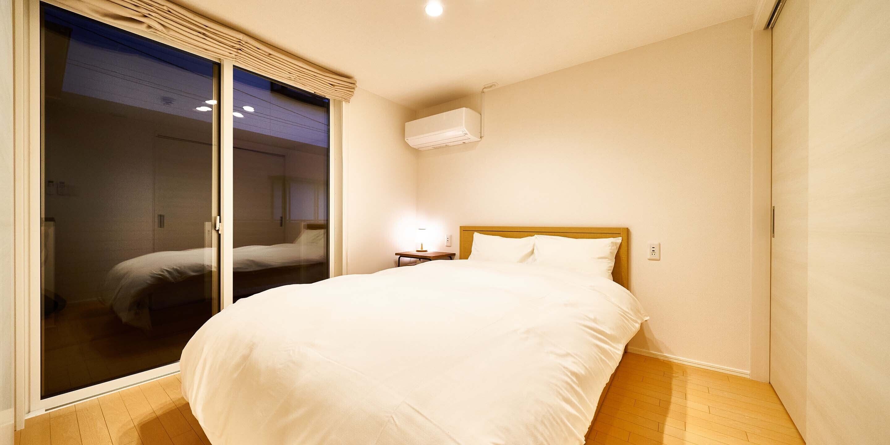 A bedroom with a large bed, a sliding glass door, a bedside table with a lamp, and an air conditioning unit on the wall.