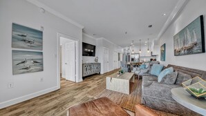 Ein geräumiges Wohnzimmer mit grauem Sofa, braunem Ledersitzhocker und einem Couchtisch. Im Hintergrund ist eine Küchenzeile mit Esstisch und Stühlen zu sehen.