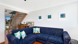 A modern living room with a blue sectional sofa, a wooden staircase, and framed artwork on the wall. - Cool View - Views of Seven Mile Beach (Gerroa)