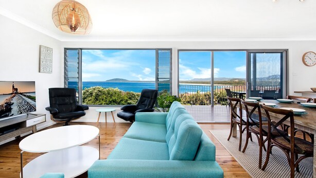 A modern living room with a large sofa, two armchairs, a dining table with chairs, and a view of the ocean through large windows. - Cool View - Views of Seven Mile Beach (Gerroa)