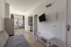 Une pièce comprenant un lit, un téléviseur fixé au mur, une petite table et un coin cuisine à l’arrière-plan.