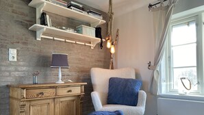 Una stanza accogliente con parete in mattoni, un mobiletto in legno, una poltrona bianca con cuscino blu e una finestra con tende.