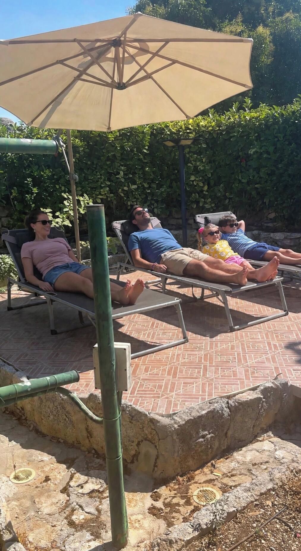 Un espace patio avec des chaises longues disposées sous un grand parasol, le tout entouré de verdure.