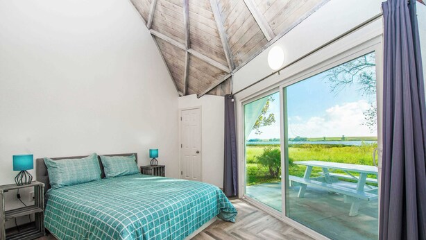 A bedroom with a large window, a bed, a nightstand, and a view of a grassy field. - STUDIO 05 - Lake Sumner (Clermont)