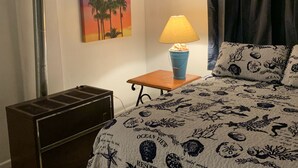 Habitación de hotel con cama con ropa de cama temática de océano, una mesita de noche con lámpara, un cuadro de un atardecer y un radiador.