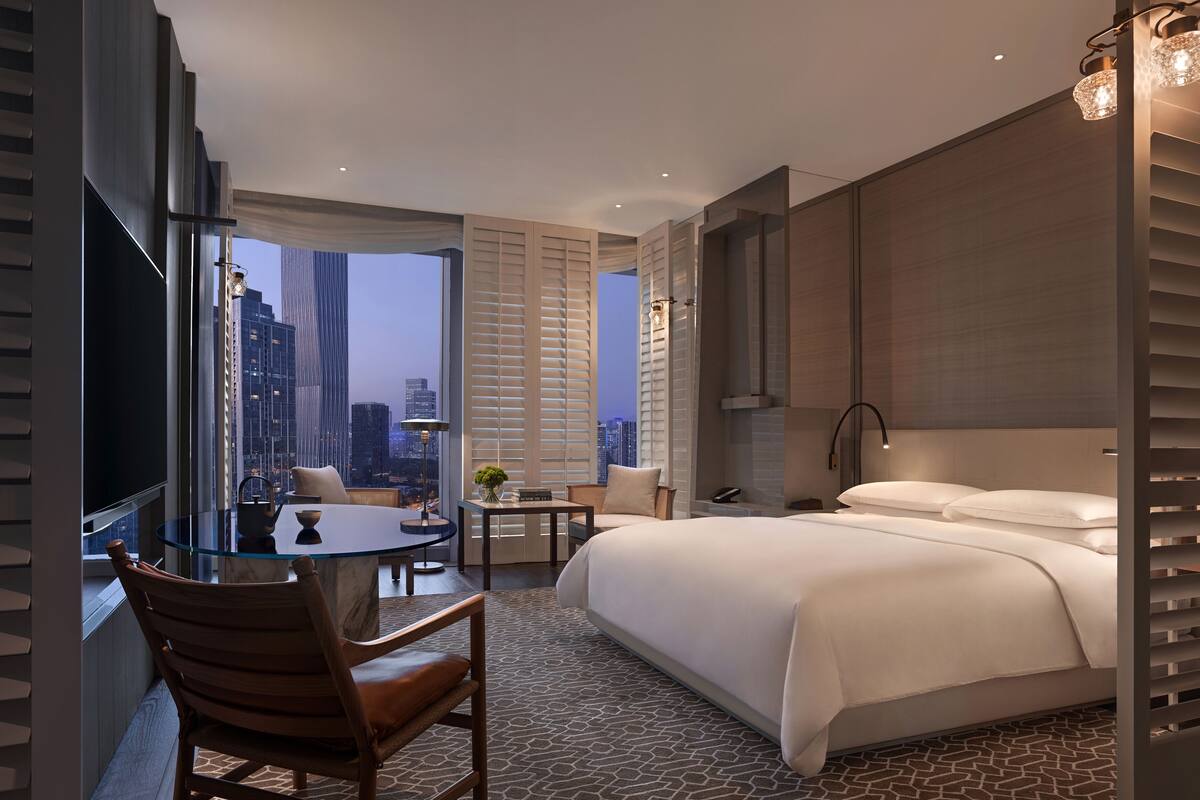 A modern hotel room with a large bed, a sitting area with a table and chairs, a TV, and a city view through large windows.