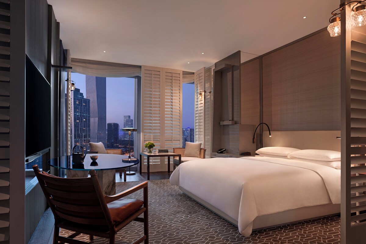 A modern hotel room with a large bed, a sitting area with a table and chairs, a TV, and a city view through large windows.