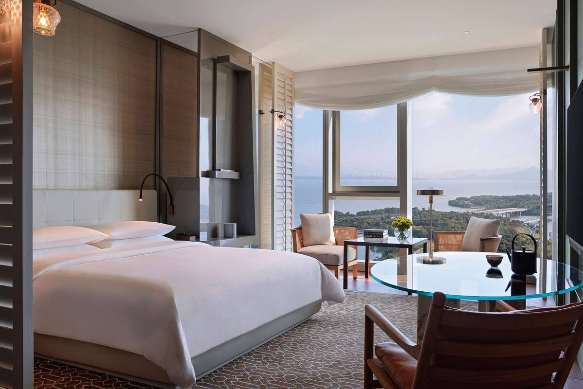 A hotel room with a large bed, a glass table, and a view of a bridge and water.