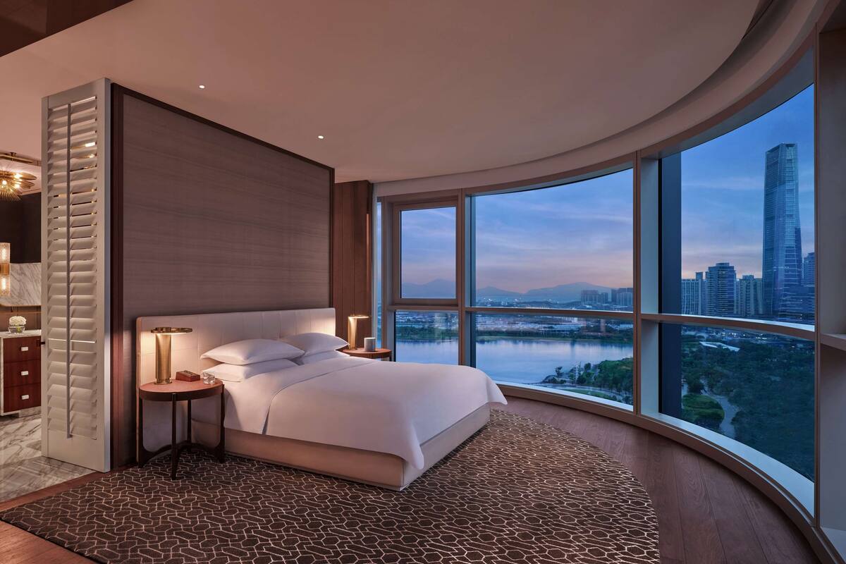 A modern bedroom with a curved window offering a city view, a large bed, a nightstand, and a mirror with lighting.