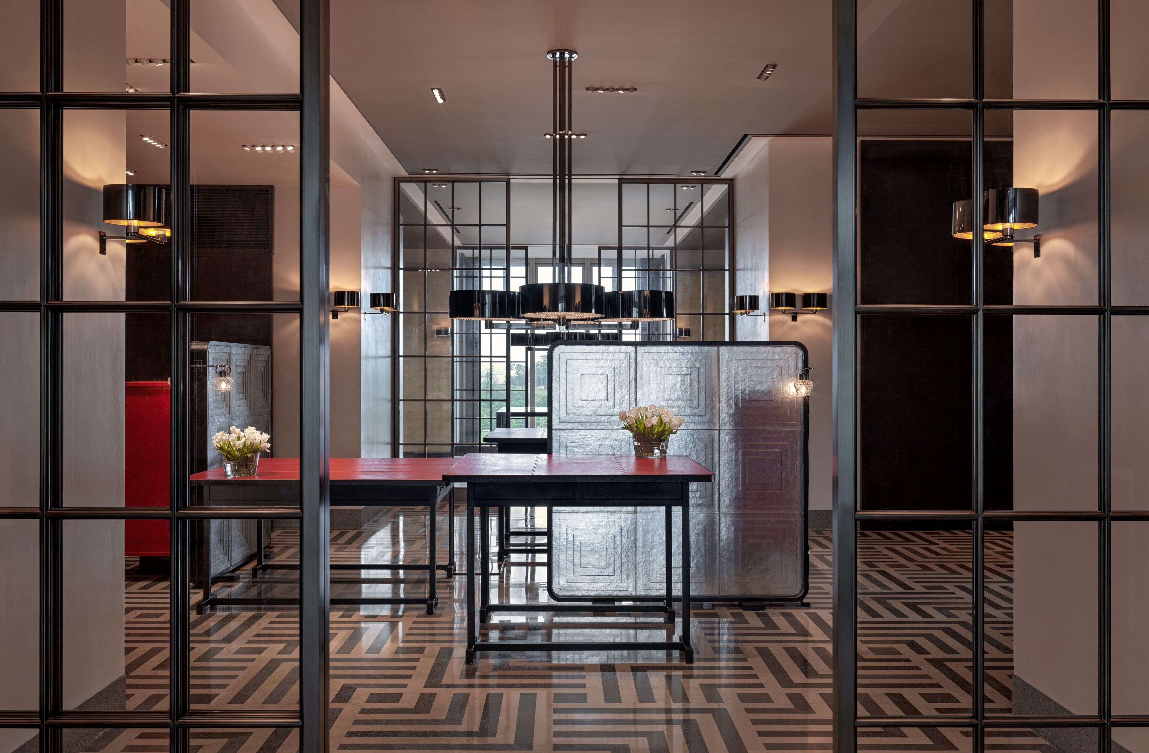 A modern hotel lobby with a geometric patterned floor, a central seating area with a table and chairs, and a decorative vase with flowers.