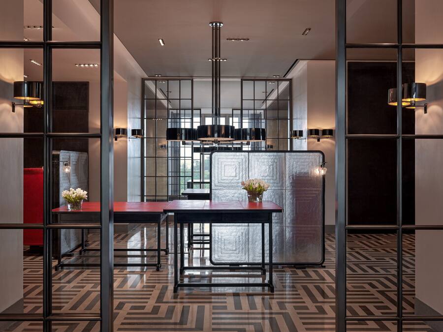 A modern hotel lobby with a geometric patterned floor, a central seating area with a table and chairs, and a decorative vase with flowers.