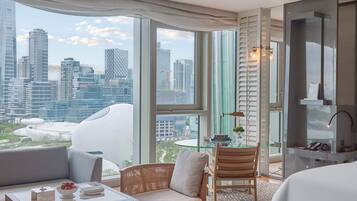 Habitación de hotel con un ventanal grande que ofrece vistas a la ciudad, una cama, un área de descanso con una mesa y un escritorio pequeño con una lámpara.