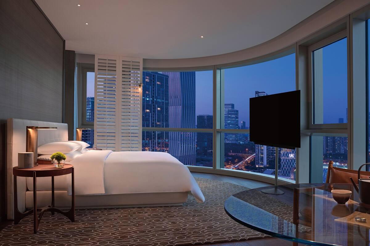 A modern hotel room with a curved glass wall, a large bed, a glass table, and a city view.