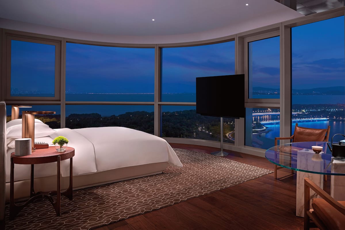 A modern bedroom with a large bed, a glass table, and a view of a cityscape at dusk.