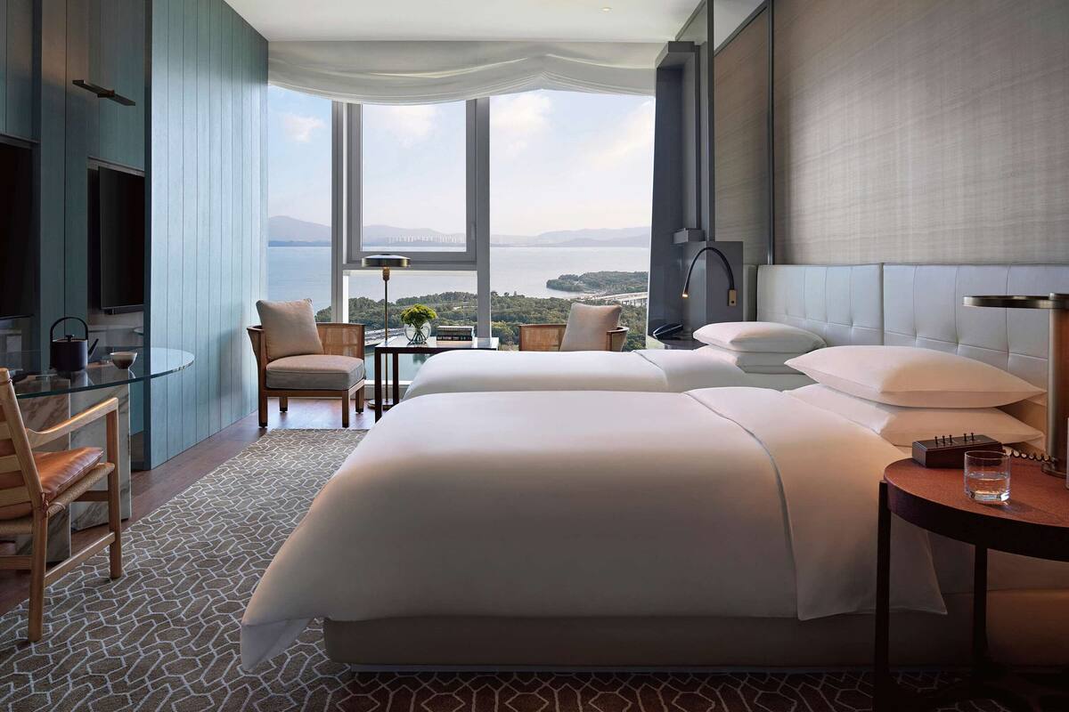 A modern hotel room with a large bed, two armchairs, a small table, and a TV. The room has large windows offering a view of the sea.
