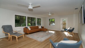 Amplia sala de estar con piso de madera, un sofá modular, dos sillones, una mesa de centro y ventanas grandes con vistas a zonas verdes.