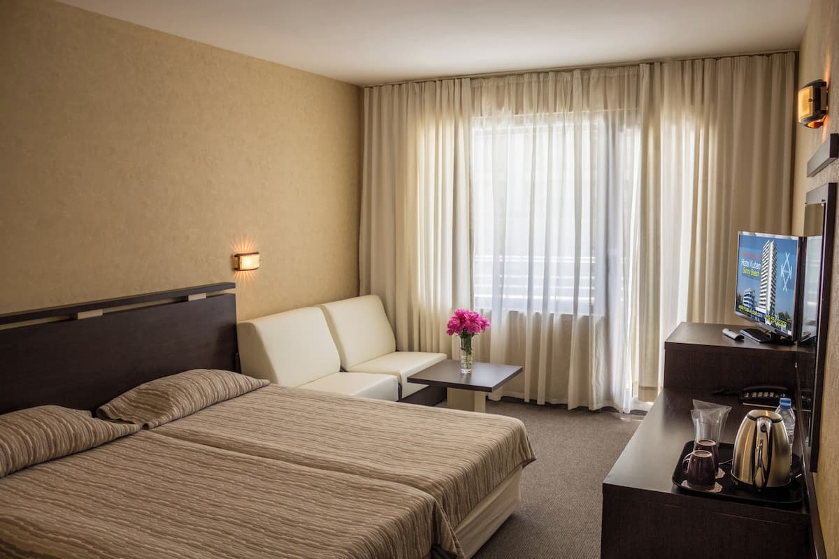 Een hotelkamer met een bed, een slaapbank, een kleintje tafeltje met een bloevas, een bureau met een televisie, en een raam met gordijnen.