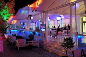 An outdoor seating area with tables and chairs, a bar, and a neon sign. - Kuban Resort and Aquapark (Nessebar)