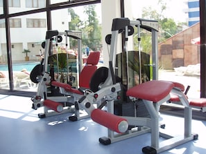 Uma academia com equipamentos de ginástica, piscina e vista para um edifício e árvores.