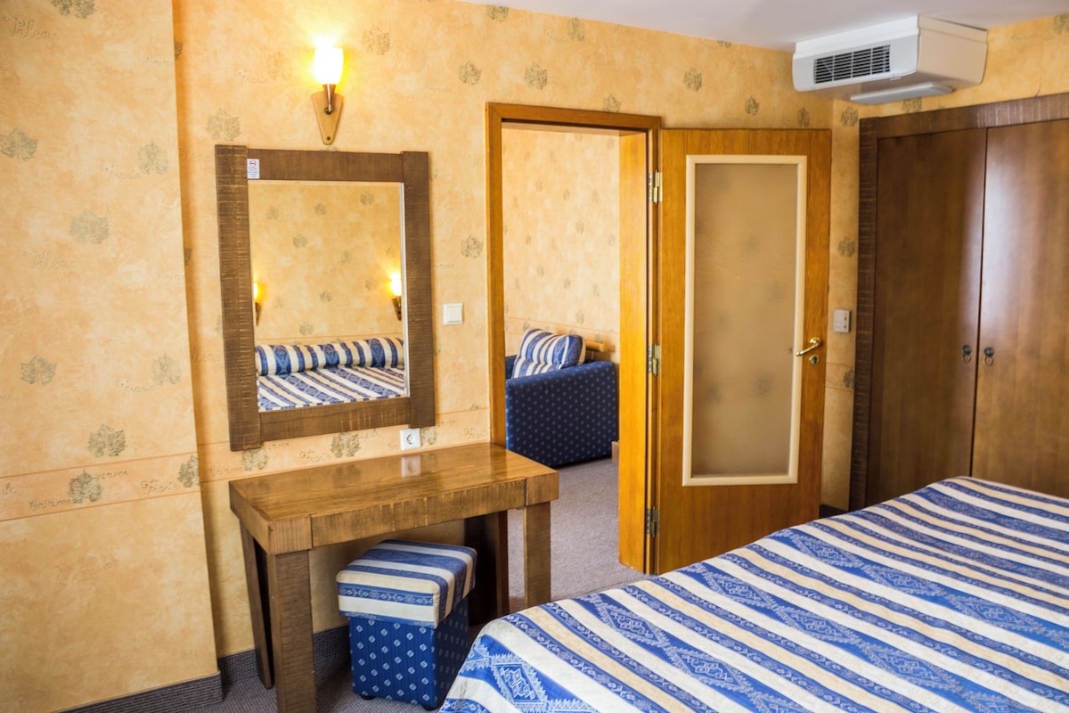 Une chambre d’hôtel comprenant un lit, un bureau en bois, un miroir, une lampe fixée au mur et une porte donnant sur une autre pièce.