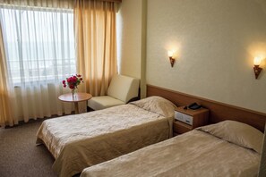 Habitación de hotel con dos camas, una mesita con un florero, una silla, mesitas de noche con lámparas y una ventana con cortinas.