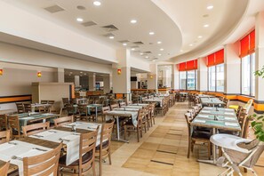 Un restaurant spacieux, doté de nombreuses tables et chaises, d’un espace cuisine et de grandes fenêtres.