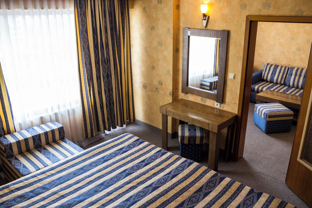 Quarto de hotel com cama, escrivaninha, cadeira, espelho e janela com cortinas.