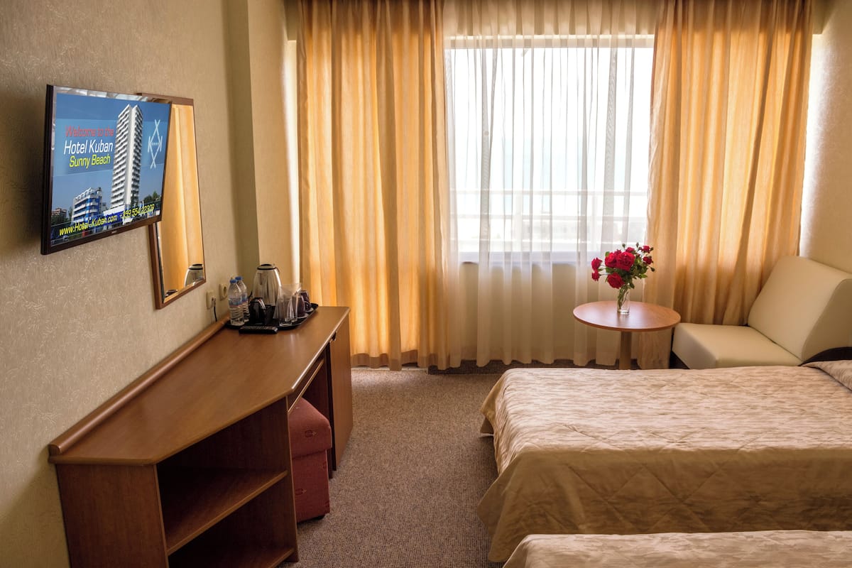 Een hotelkamer met een bed, een bureau met een computerscherm, een kleine tafel met een bloemenvaas en een raam met gordijnen.