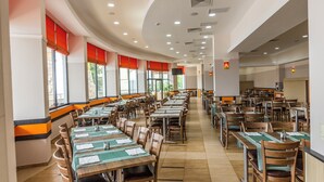 Un vaste espace dédié à la salle à manger, avec de nombreuses tables et chaises, de grandes fenêtres équipées de stores rouges et un plafond incurvé doté d’un éclairage encastré.
