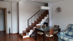 Una escalera con peldaños de madera y barandales negros, una mesa de comedor de vidrio con sillas, y un sofá a cuadros azul y amarillo.