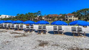 A beach with rows of blue umbrellas and beach chairs, a large building in the background, and a clear blue sky. - New Inventory! Updated Kitchens and Baths 2 BR Bikes & Beach Gear included (Hilton Head Island)