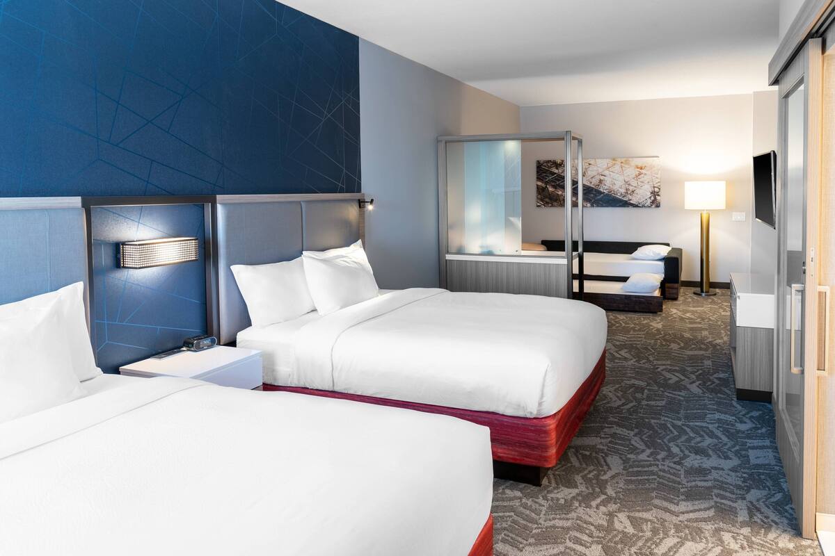 A hotel room with two beds, a bedside table, a lamp, a TV, and a large window.