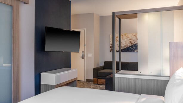 A modern hotel room with a flat-screen TV mounted on the wall, a small seating area with a chair, a bedside table, and a large window with a view of the interior.