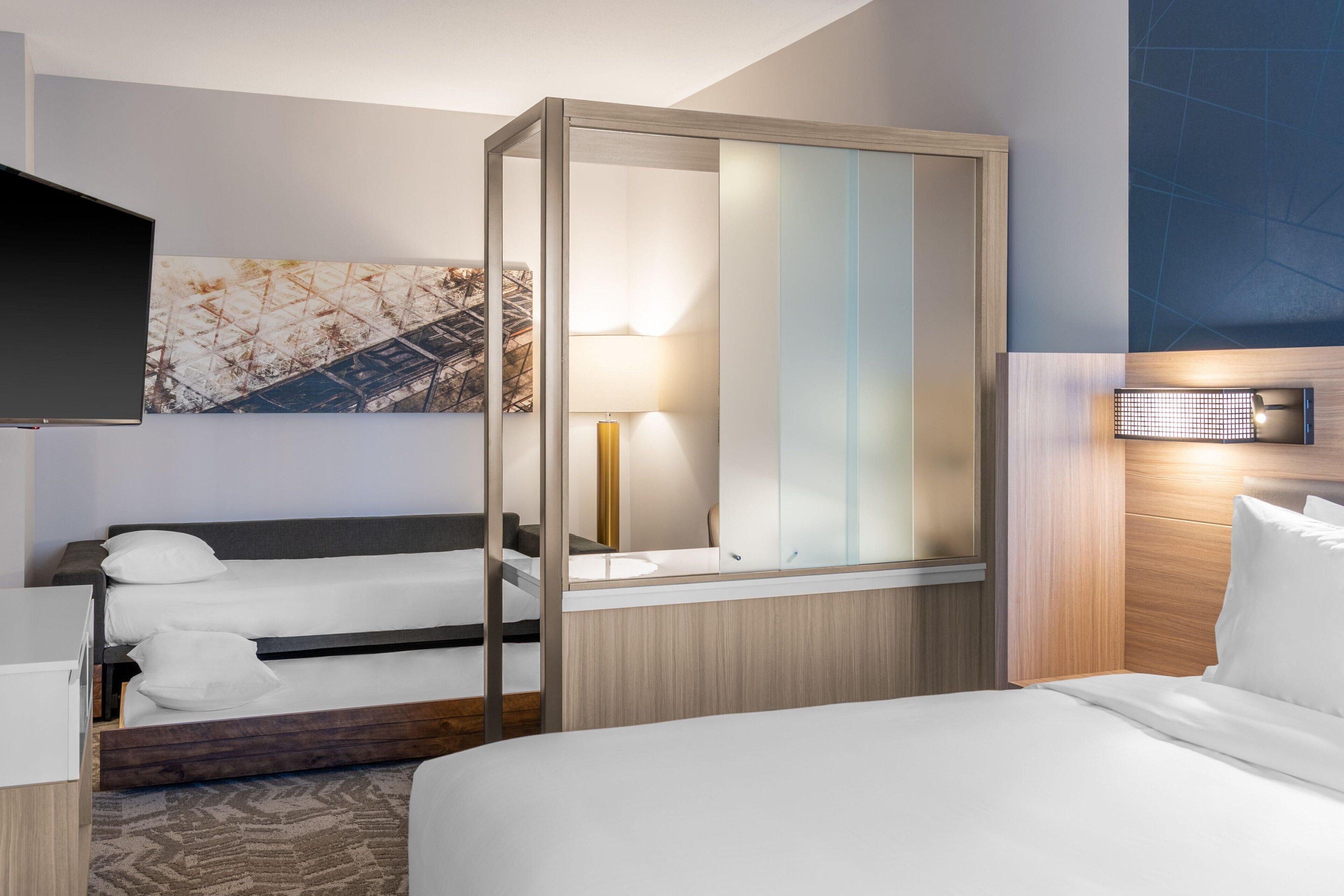 Une chambre d’hôtel équipée d’un lit, d’un bureau, d’une chaise, d’une télévision, d’un miroir et d’une lampe fixée au mur.