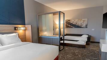 Quarto de hotel com uma cama grande, uma cama menor, uma escrivaninha com abajur e um espelho amplo.