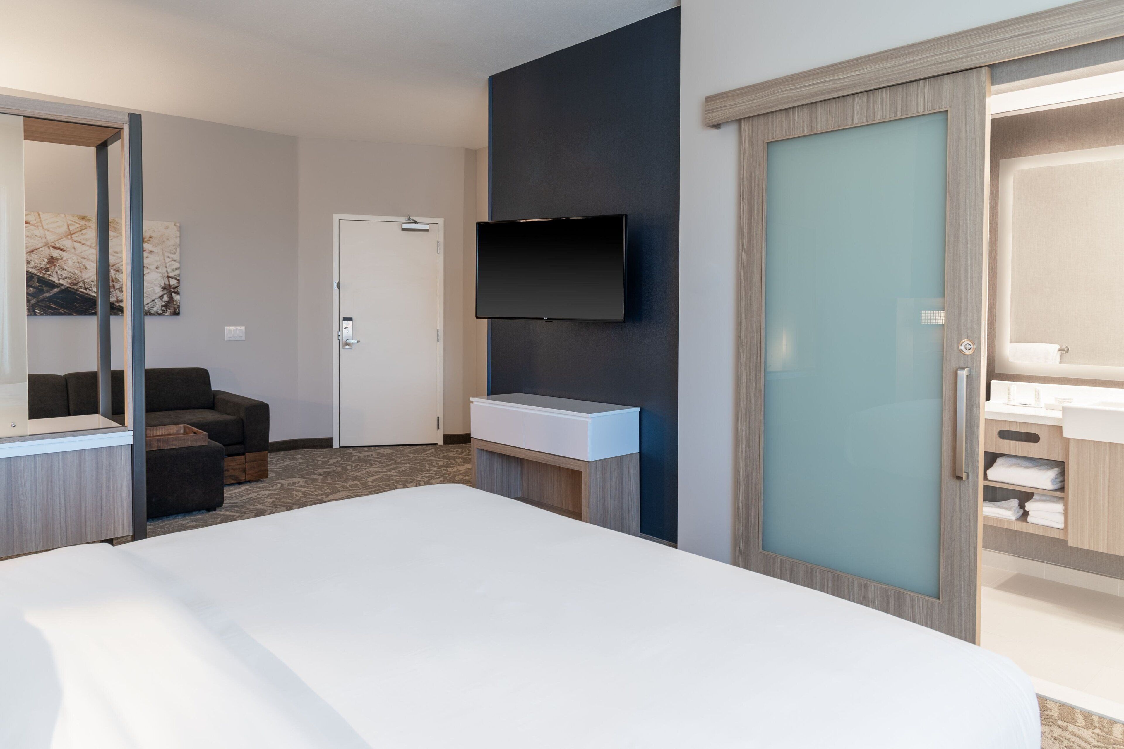 A modern hotel room with a large bed, a flat-screen TV mounted on the wall, a bedside table, and a wardrobe with a frosted glass door.
