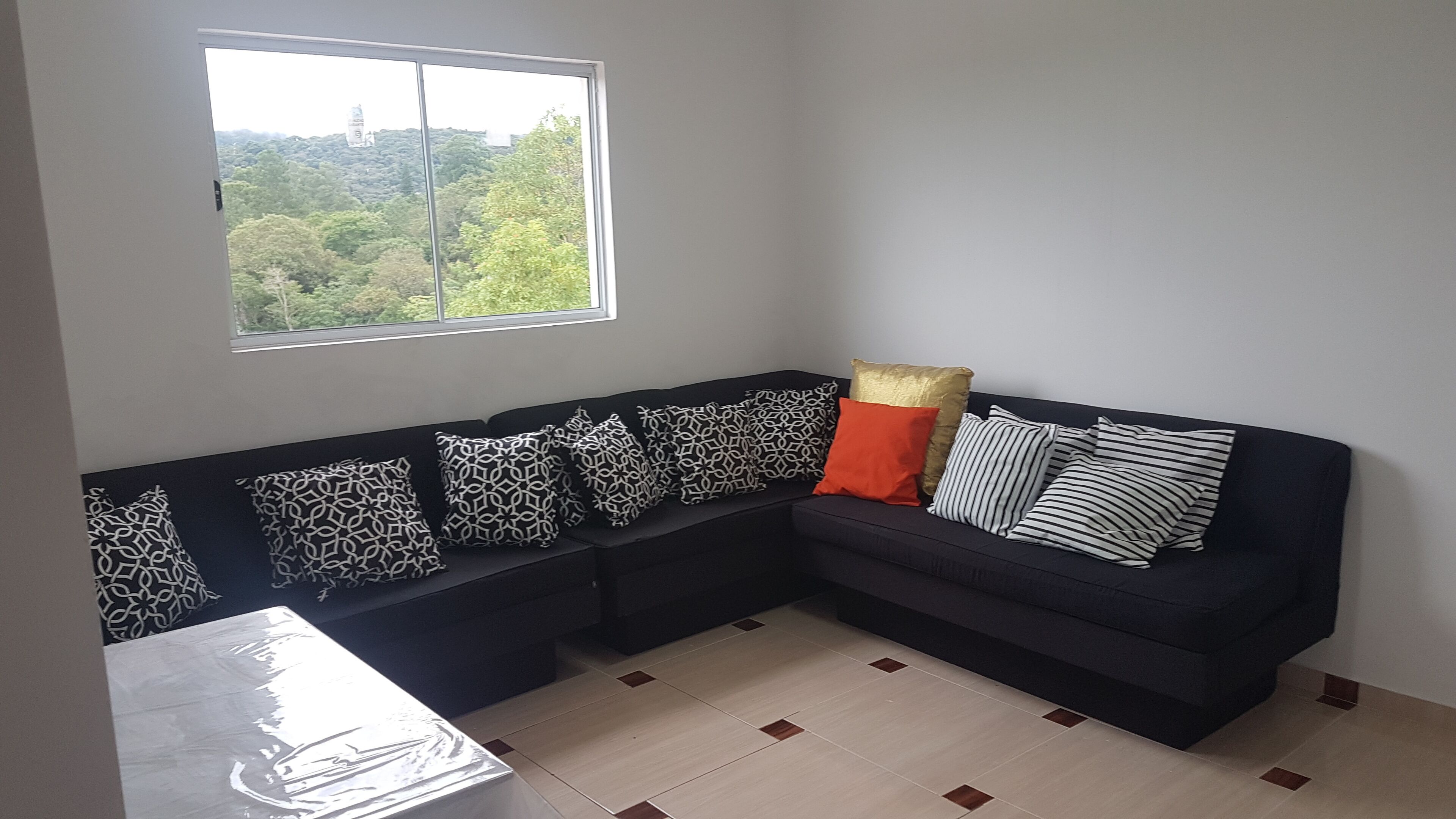 A modern living room with a black sectional sofa, patterned pillows, a small table, and a window overlooking a forested area.
