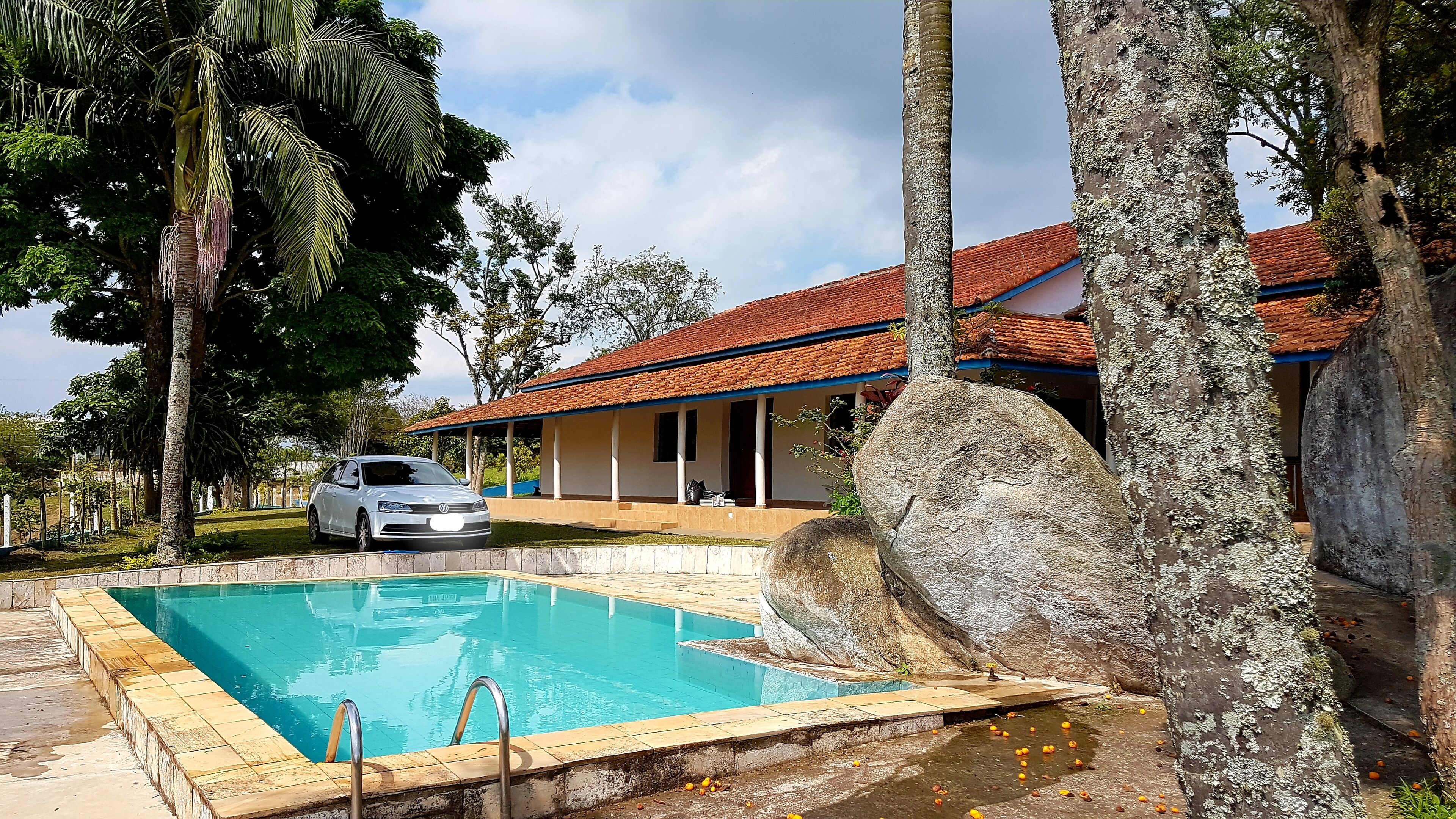 Una piscina con un coche aparcado cerca, un edificio con tejado rojo y rocas grandes alrededor de la zona de la piscina.