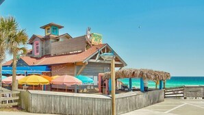 A beachside building with colorful facades, a thatched roof structure, and a wooden deck with umbrellas. - Sandy Toes Gated Community*Pool*Gym*Beach Svc! (Miramar Beach)