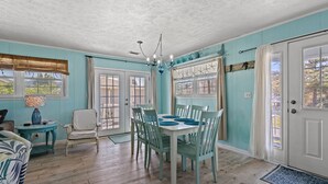 Un coin repas avec une table blanche et des chaises bleues, un lustre et une vue par la fenêtre.